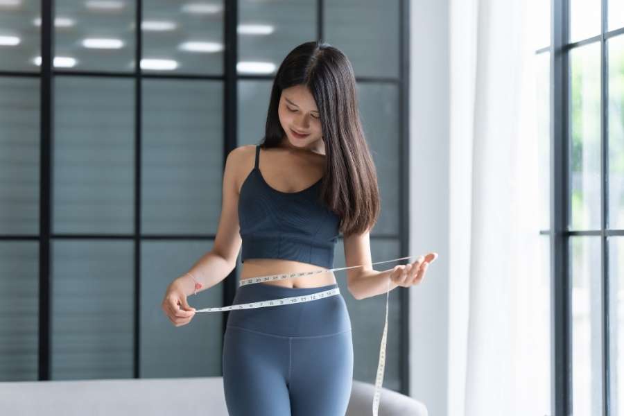 Une femme en tenue de sport mesure son tour de taille avec un mètre ruban pour suivre sa progression pour perdre du poids.