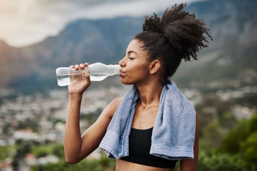 Une femme en tenue de sport, une serviette sur les épaules, boit de l'eau fraîche pour purifier son organisme en profondeur après l'effort.
